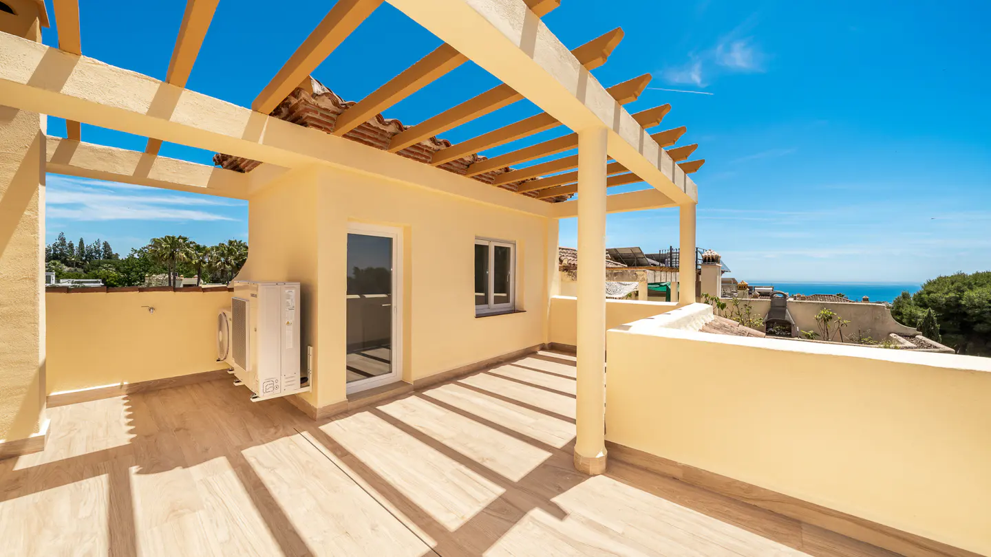 A sunny balcony with a pergola casts shadows on the light-colored tile floor. A door and window are visible, with a view of the ocean in the background.