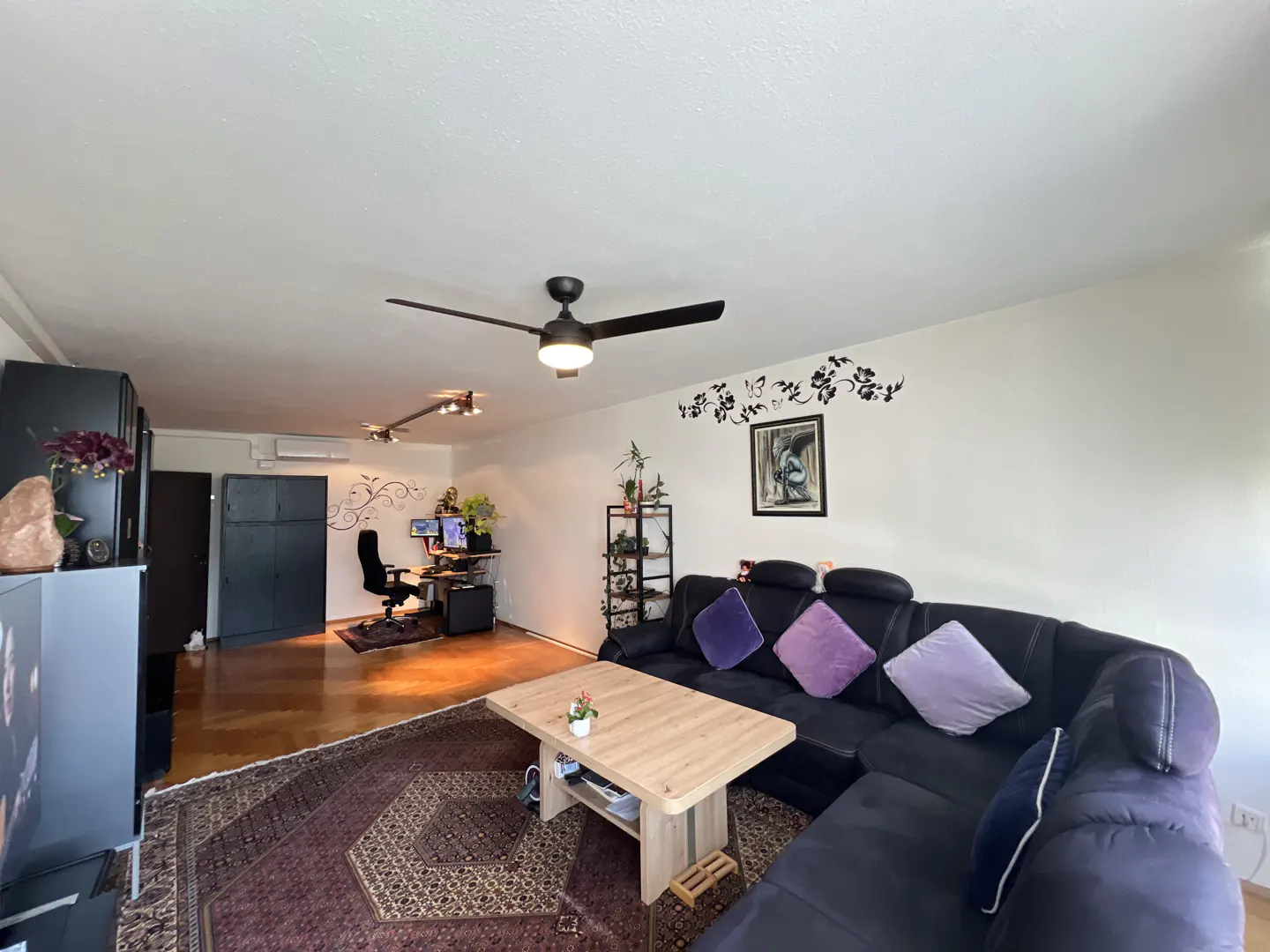 Spacious living room with a black sectional sofa, wooden coffee table, patterned rug, and a home office area in the back.