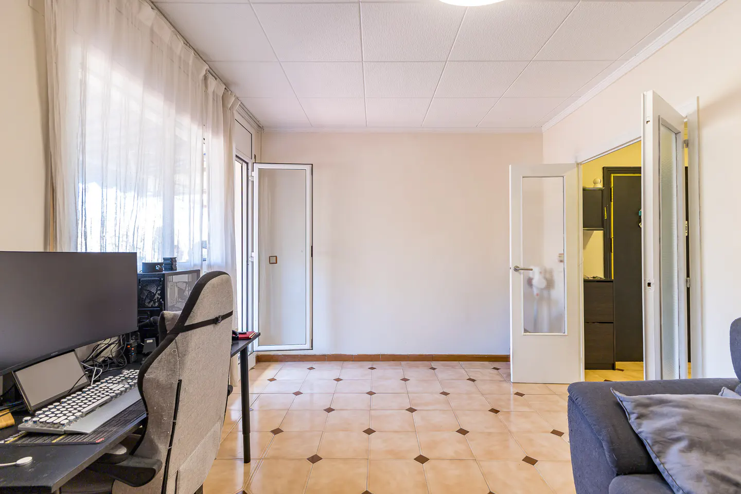 Bright room with beige walls, tiled floor, and white doors. A desk with computer and chair are on the left, and a blue sofa is on the right.