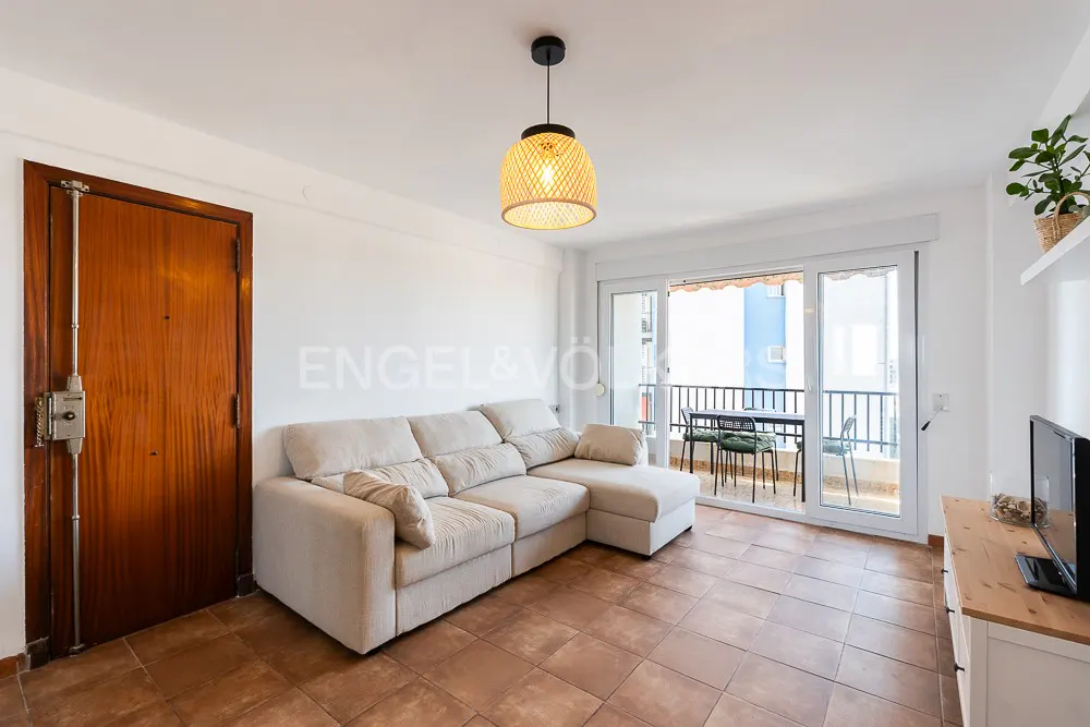 Living room with a beige sofa, brown tiled floor, and a balcony with a table and chairs. A wooden door is on the left.