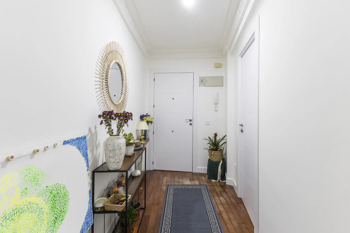 Bright hallway with white walls, wood floors, and a console table. A round mirror hangs above flowers and plants. A blue rug leads to a white door.