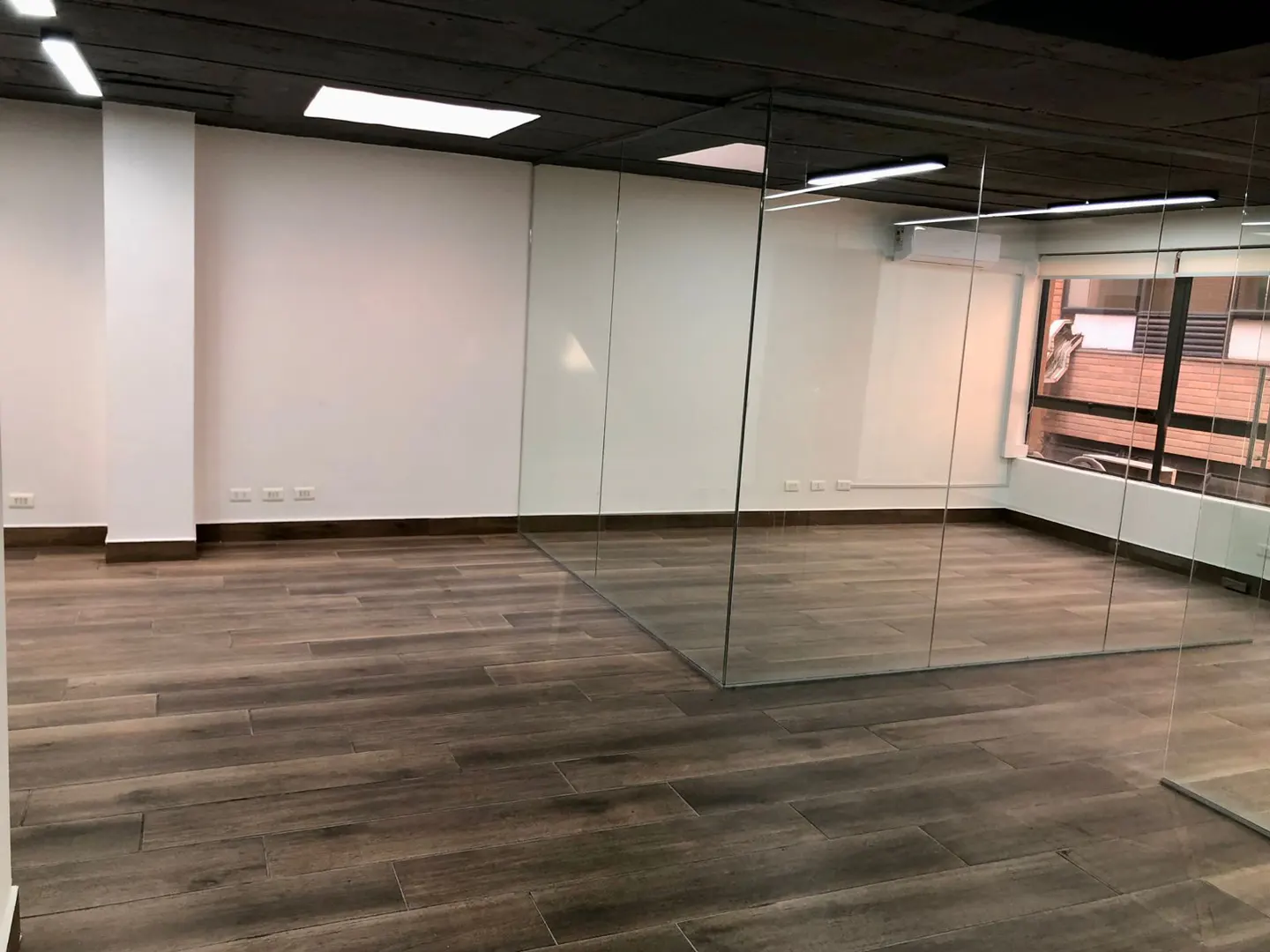 An empty office space with wood-look tile floors, white walls, and a black ceiling. Glass partitions divide the room, and a window shows a brick building outside.