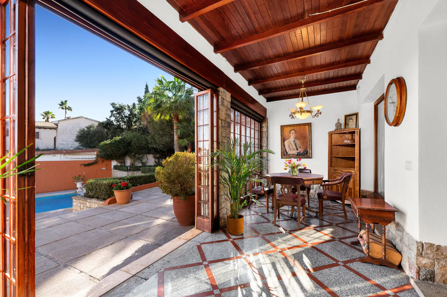 Indoor dining area with wood ceiling, table, chairs, and a view of a pool and garden through open doors.
