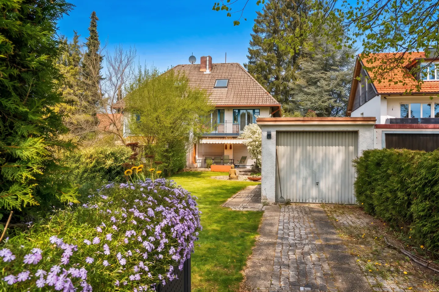 A two-story house with a brown roof and a white garage, surrounded by green trees and purple flowers.