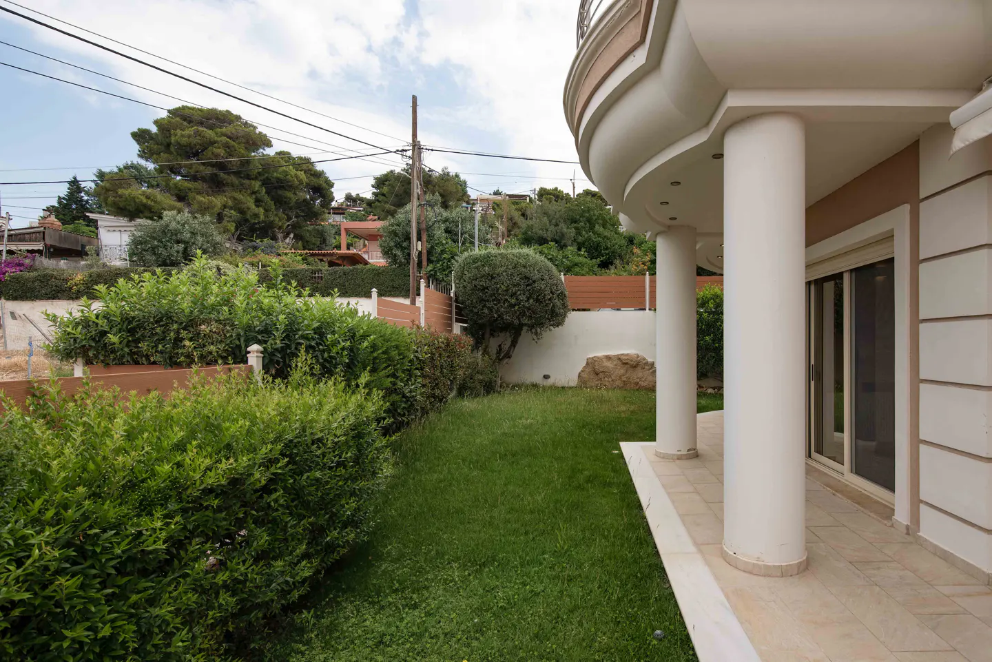 A curved house exterior with white columns, a tiled patio, and a green lawn bordered by tall hedges and trees.