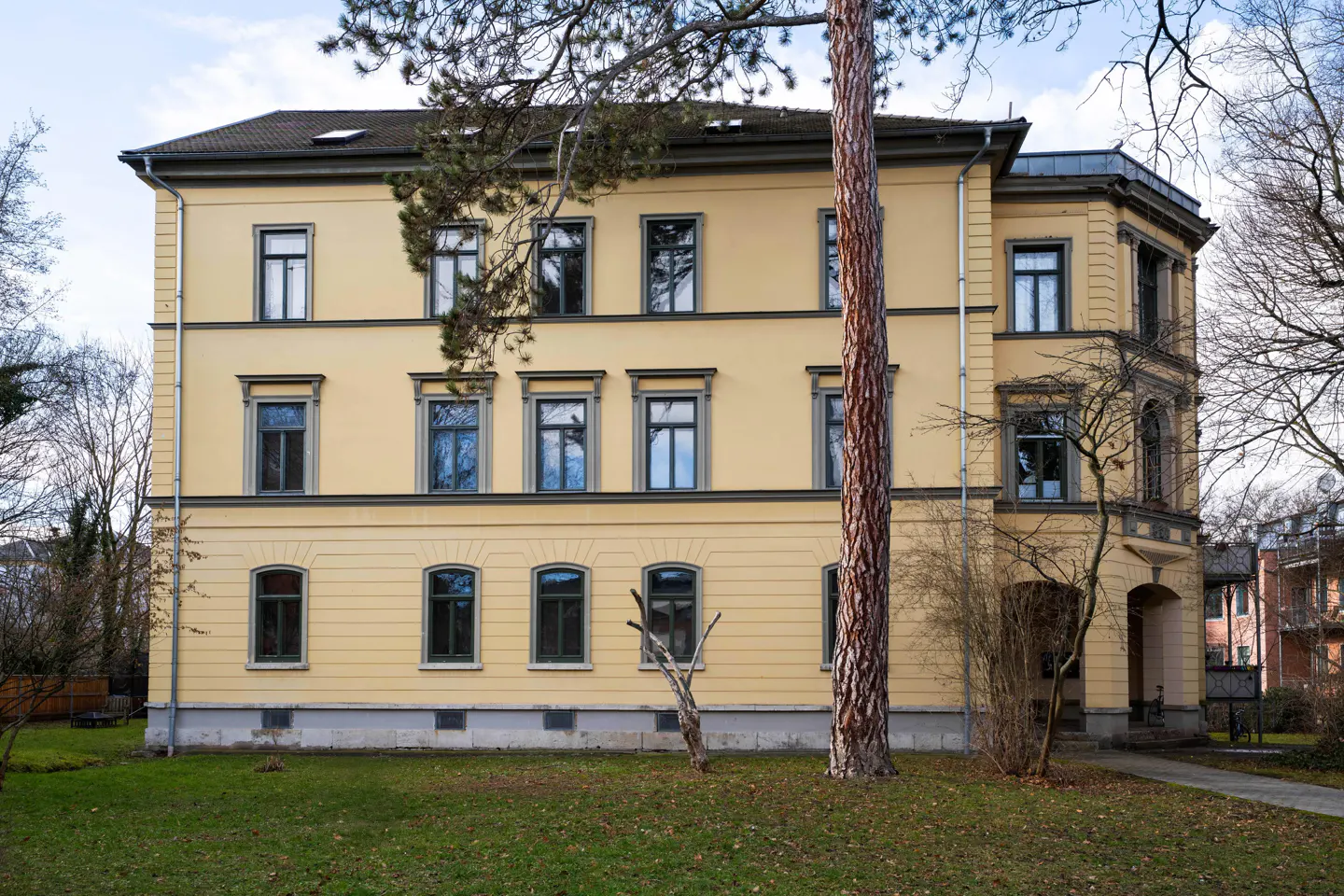 Three-story yellow building with dark trim and windows, set on a green lawn with trees.