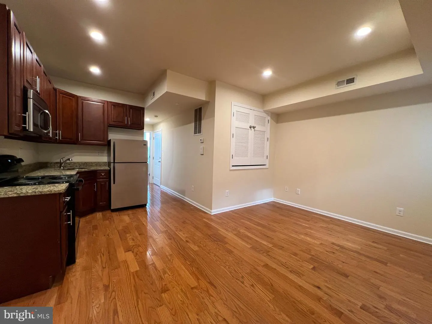 A wide shot of a kitchen and living room with hardwood floors and recessed lighting. The kitchen has dark wood cabinets and stainless steel appliances. The living room has a white shuttered window.