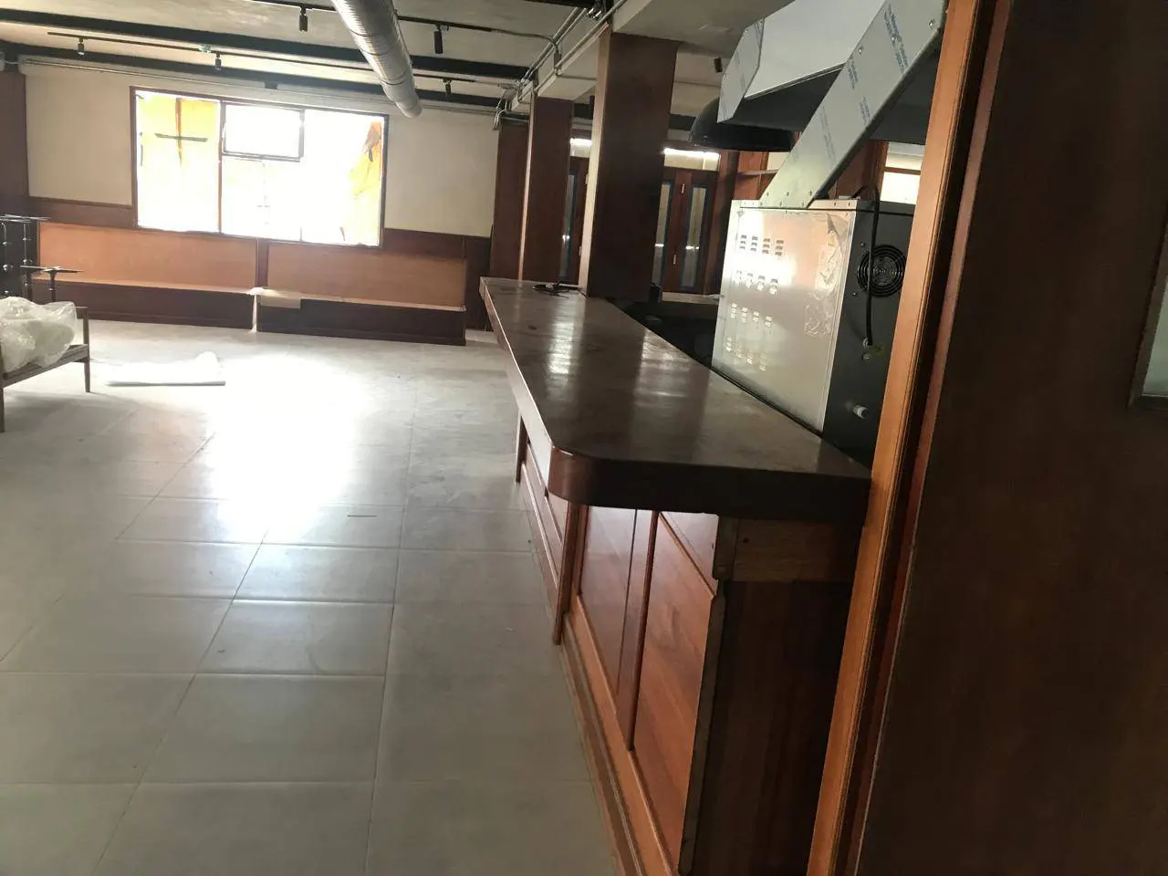 Empty restaurant space with a long, dark wood bar, stainless steel kitchen equipment, and built-in seating by a window.