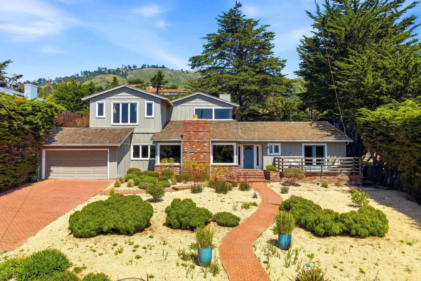Two-story gray house with a brick chimney and a blue front door. A brick path leads to the house through a sandy yard with green bushes.