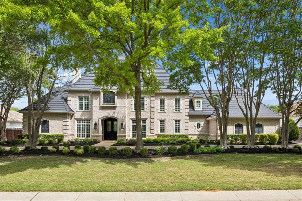 Two-story beige brick house with a gray roof, surrounded by green trees and a well-manicured lawn.