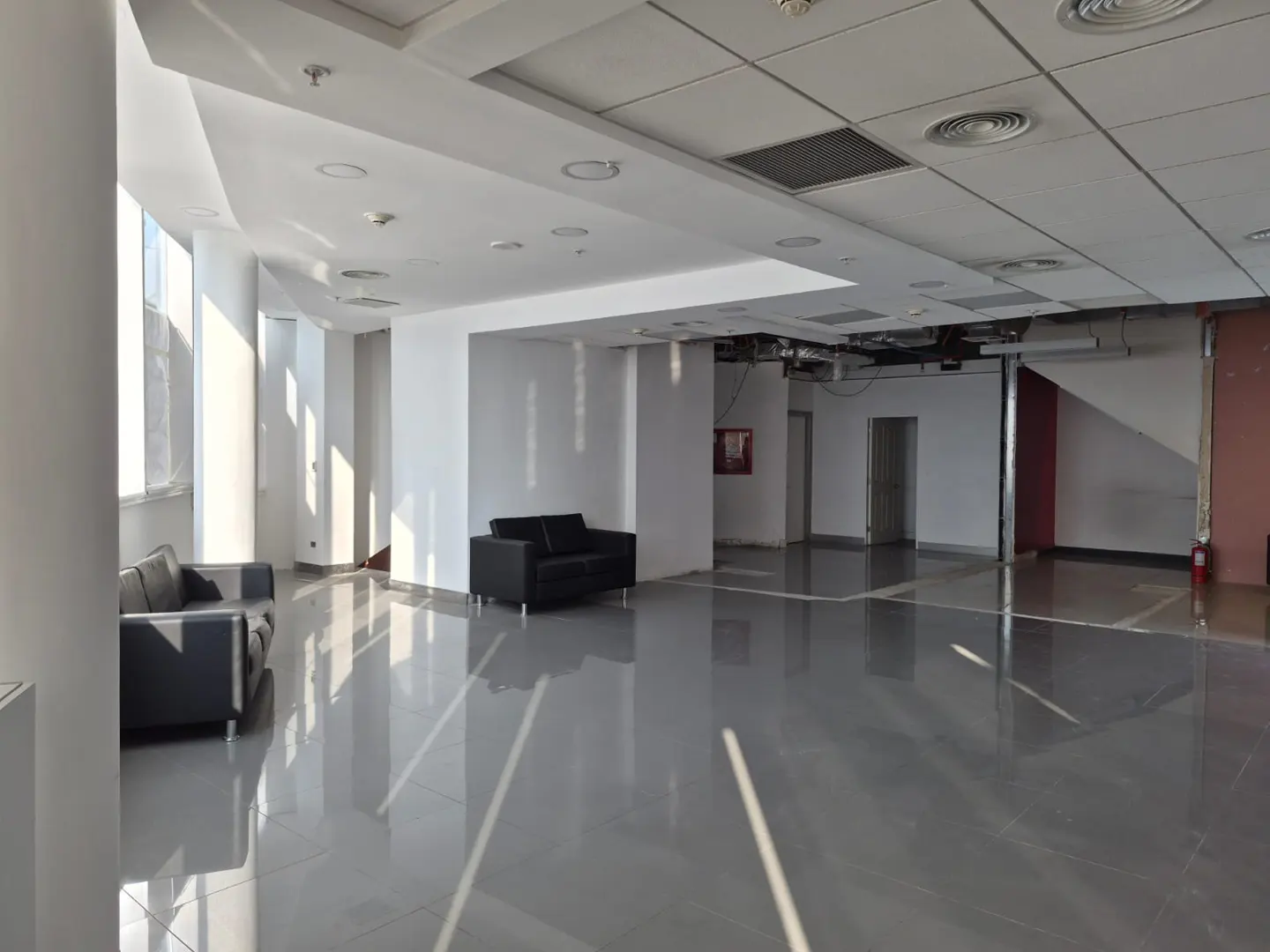 Bright, empty office space with gray floors, white walls, and black sofas. Sunlight streams through large windows.