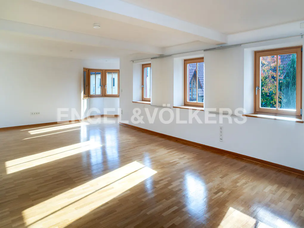 Bright, empty room with wood floors and white walls. Windows show trees and a building. Sunlight streams across the floor.