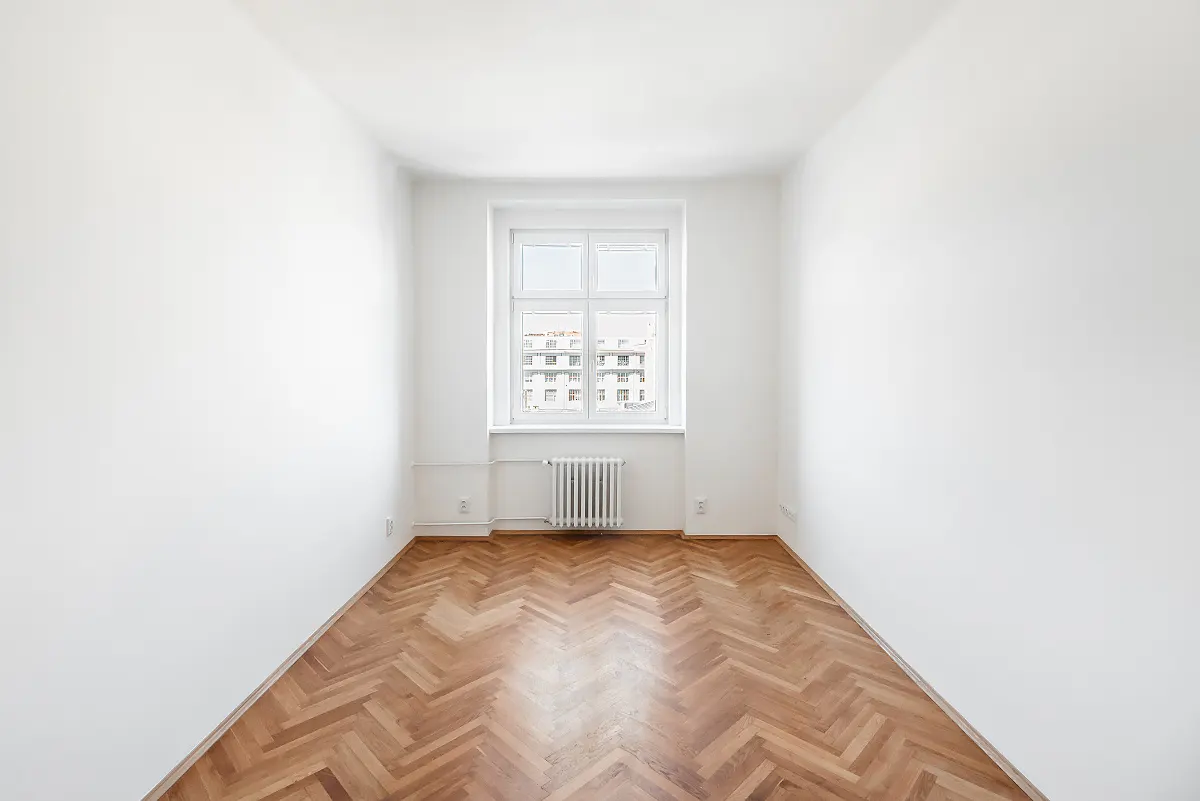 Empty room with white walls and herringbone wood floor. A window with a radiator below looks out onto a building.
