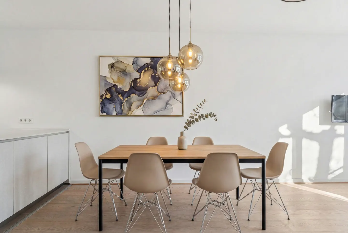 Bright dining room with a wood table, beige chairs, and a modern light fixture. An abstract painting hangs on the white wall.