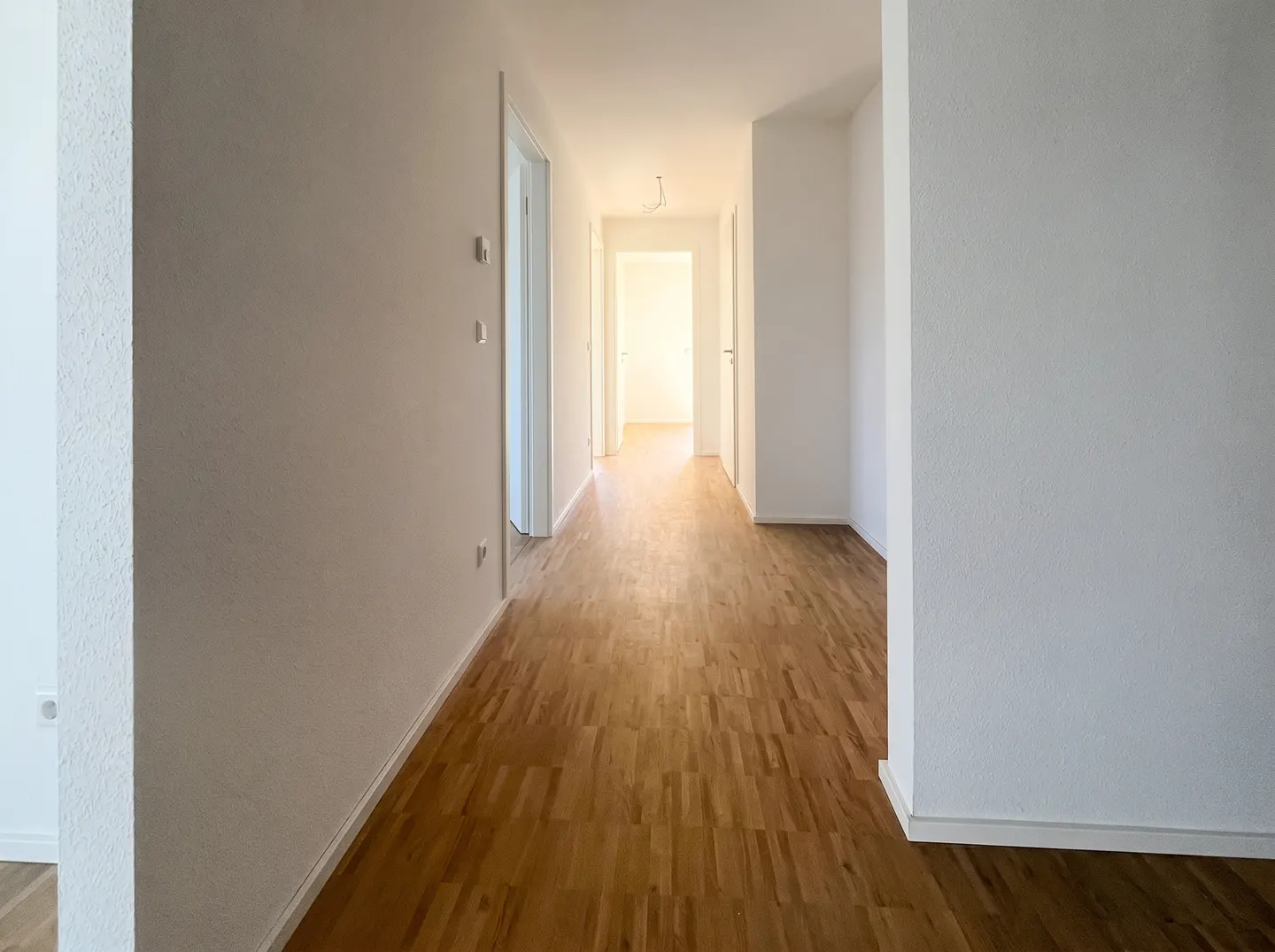 A bright hallway with white walls and a light brown parquet floor stretches into the distance.