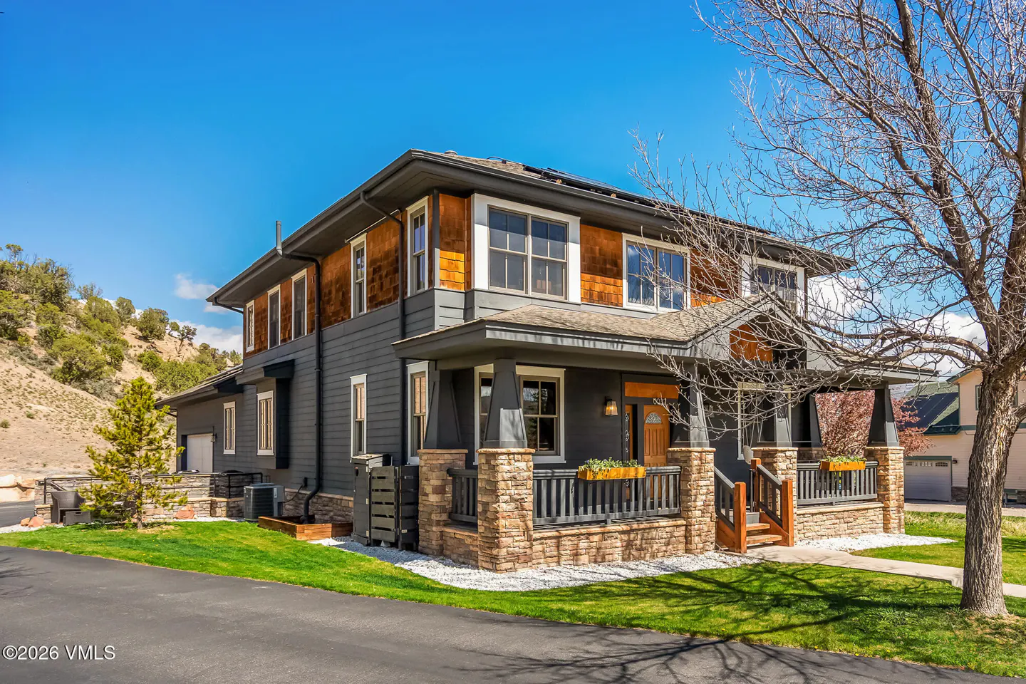 Two-story house with gray siding, brown shingles, and a stone porch under a blue sky.