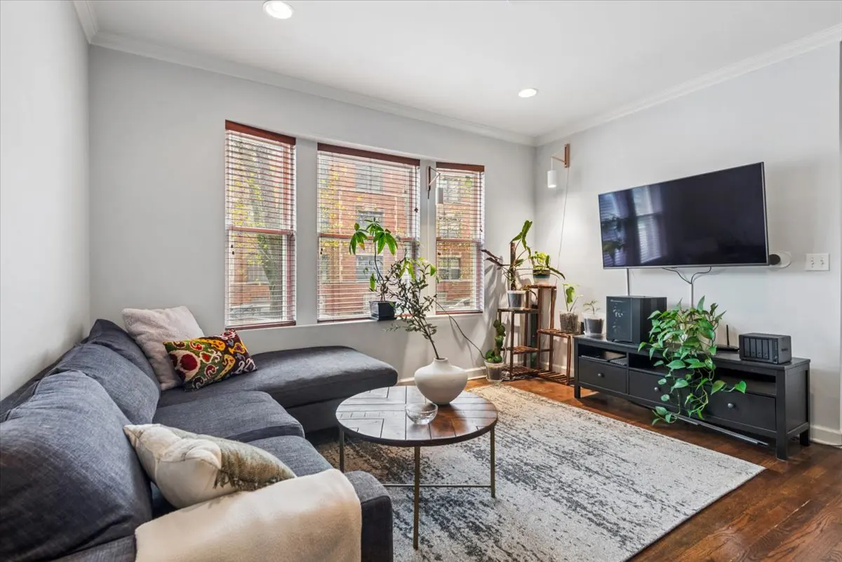 Living room with a gray sectional sofa, round wood coffee table, and a black TV stand with a flat-screen TV above it. There are plants throughout the room.