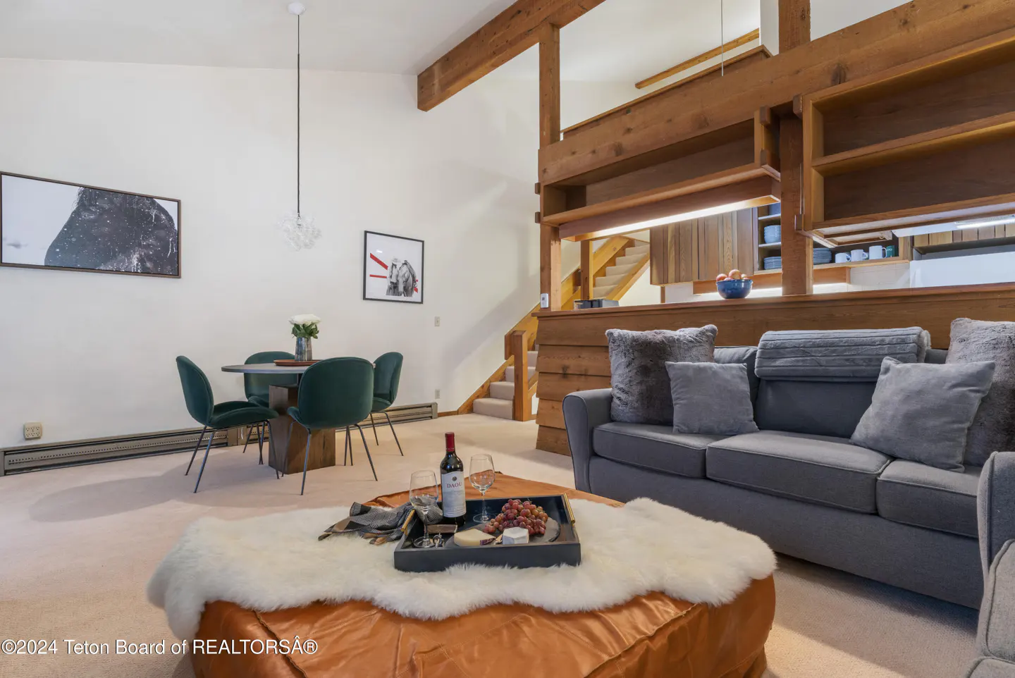 Living room with gray sofa, orange ottoman with wine and cheese, and a dining table with green chairs. Wooden beams accent the high ceiling.