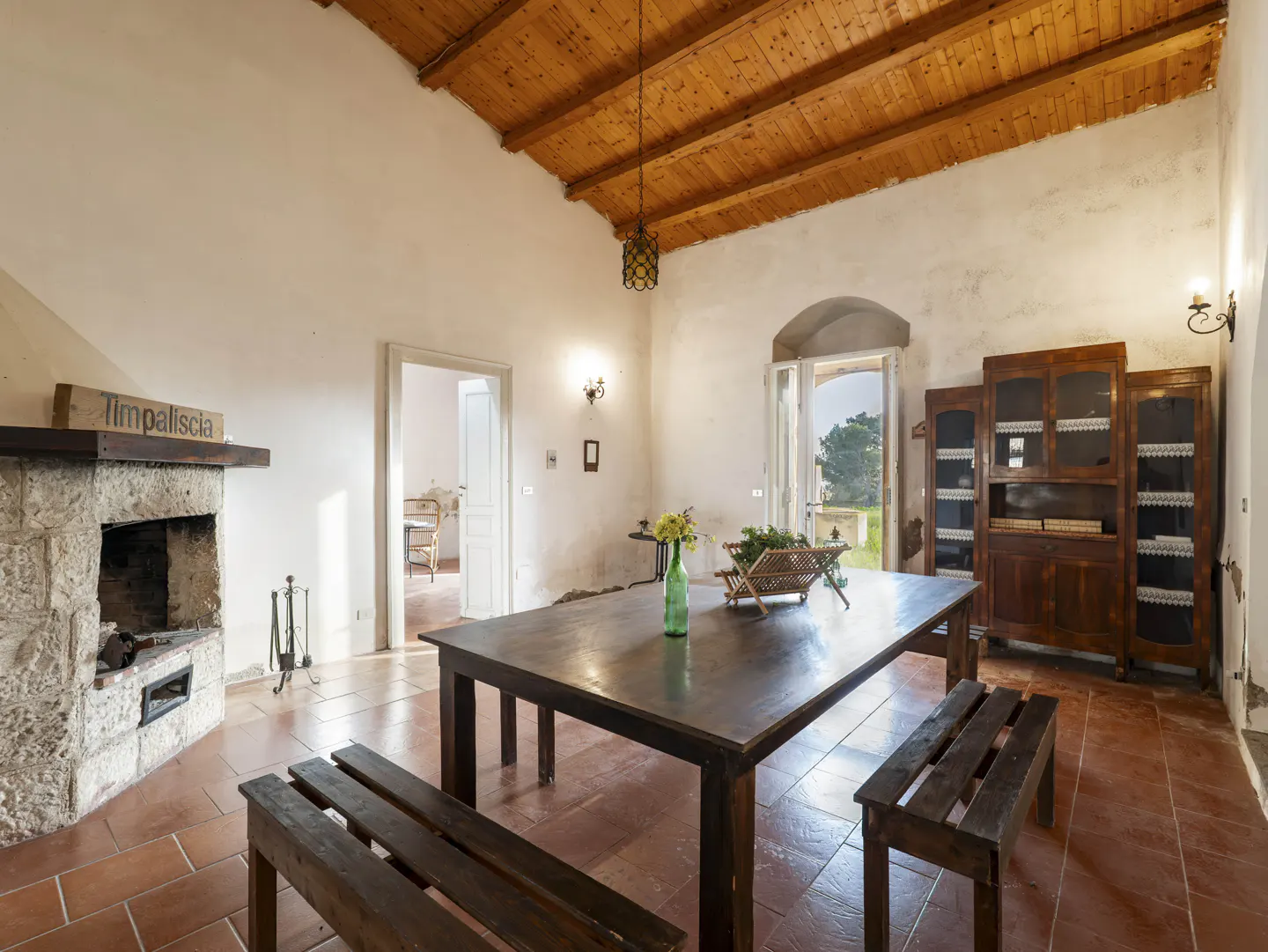 Rustic dining room with a stone fireplace, wooden table and benches, and a view to the outside.