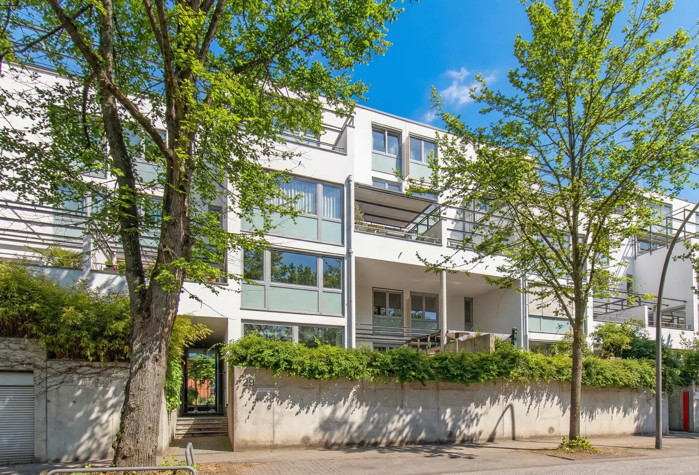 Modern white apartment building with balconies, green trees, and a concrete wall with green bushes. Blue sky.