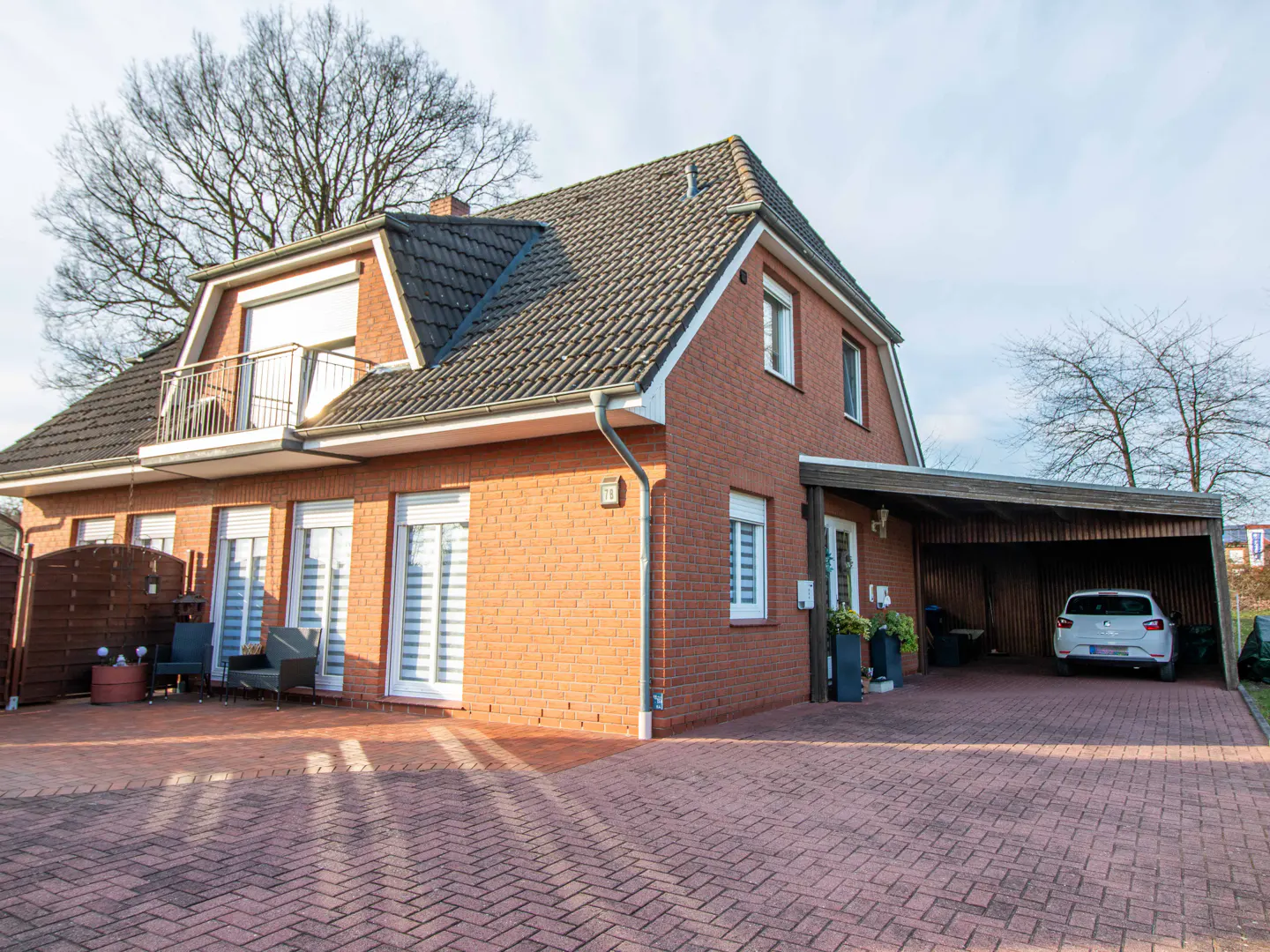 Two-story brick house with a brown roof, balcony, and carport with a white car parked inside. A brick driveway leads to the house.