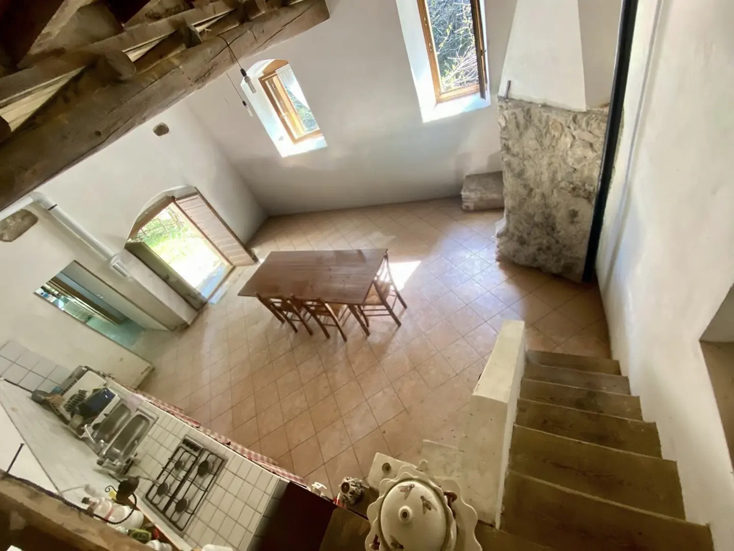Angle shot of a bright, open-plan living space with a kitchen, dining table, and stairs. Skylights illuminate the room with white walls and tiled floors.