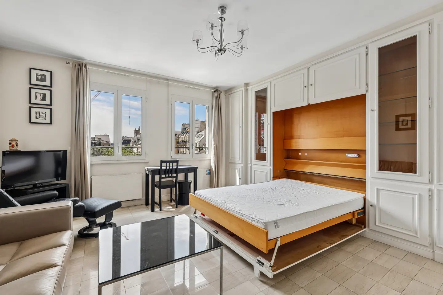 Bright living room with a pull-down bed, white cabinets, a black desk, and a leather sofa. Windows show a cityscape.