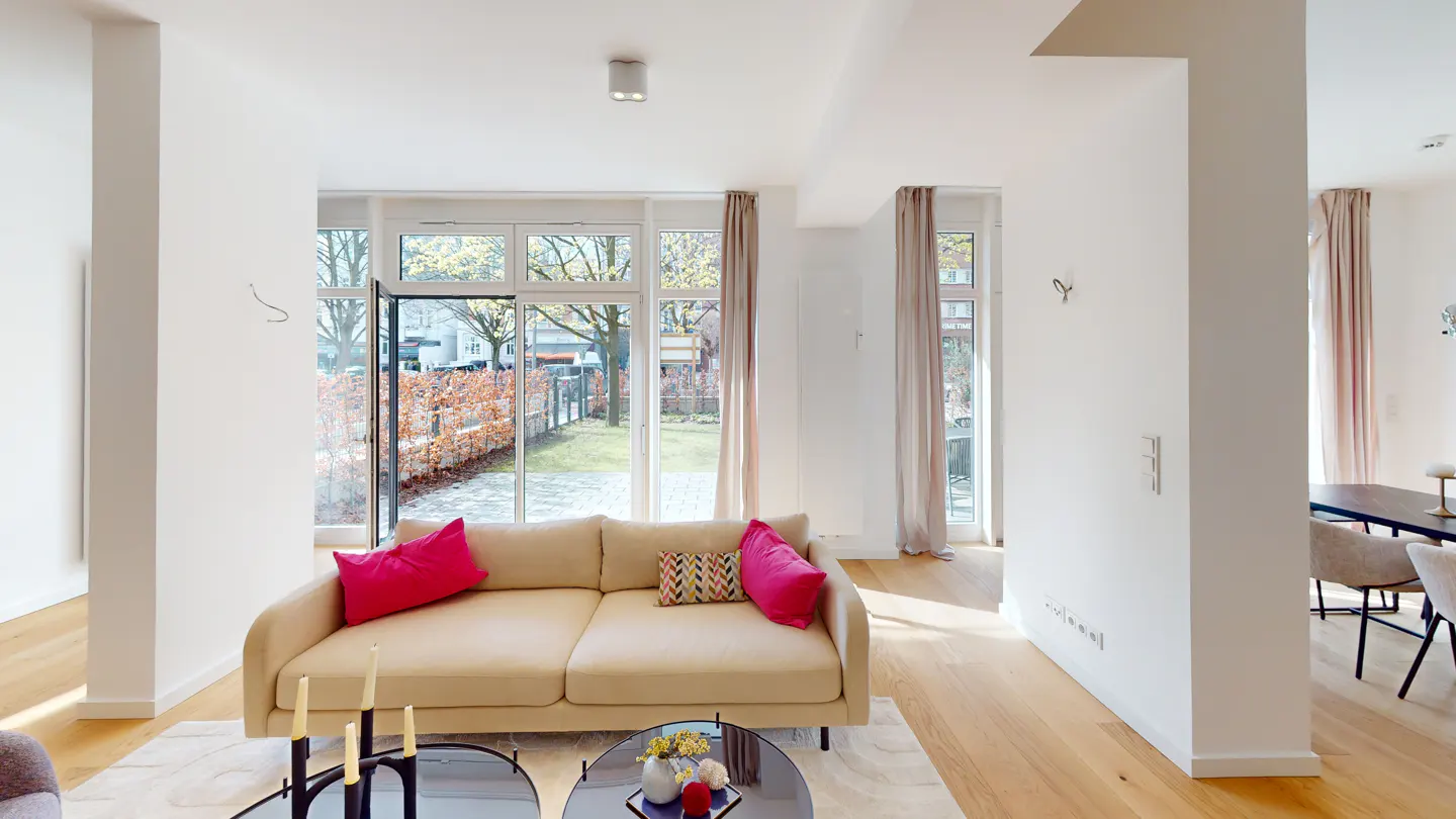 Bright living room with a beige sofa, pink pillows, and a glass coffee table. Large windows overlook a garden. Hardwood floors and white walls create a modern space.