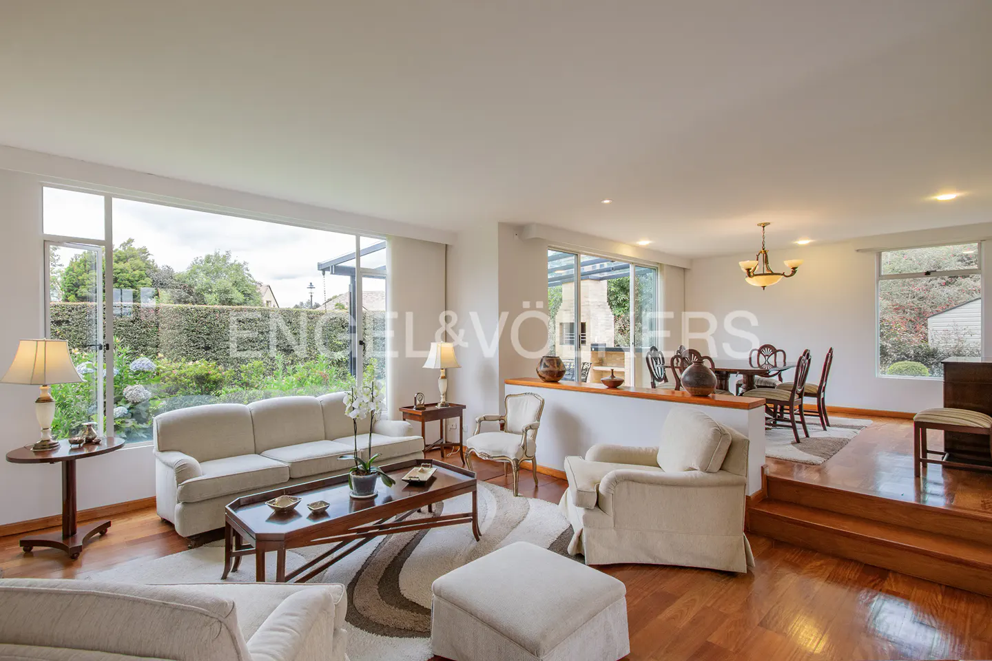 Open-concept living and dining room with hardwood floors, white sofas, and large windows overlooking a green garden.