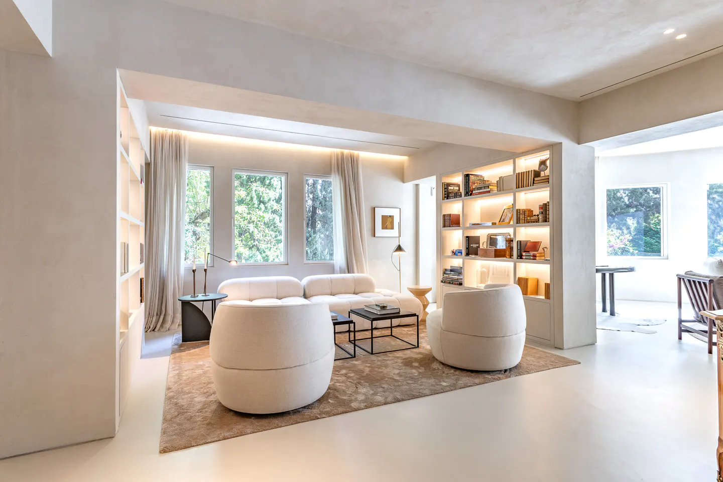 Bright, modern living room with white sofa, chairs, and built-in bookshelf with warm lighting. Windows offer a view of lush greenery.