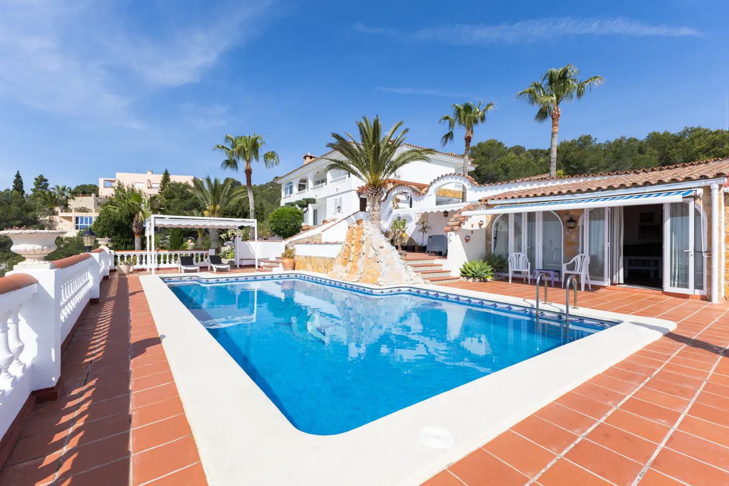 A bright blue swimming pool is surrounded by a red tile patio at a white house with palm trees under a blue sky.