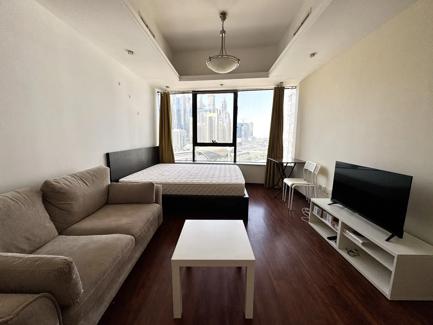 A studio apartment with a beige sofa, bed, white table, TV, and a large window with a city view.