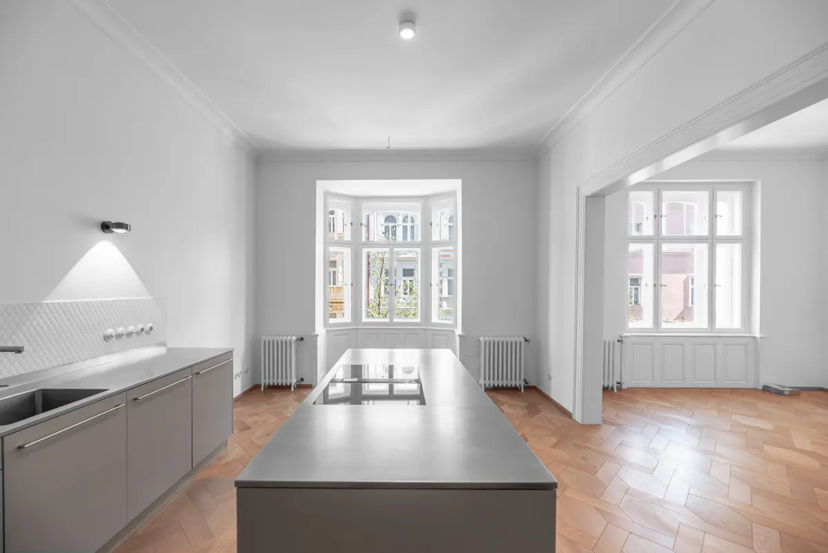 Bright, empty kitchen with white walls, herringbone wood floor, and gray cabinets. A large island sits in the center, with a bay window in the background.