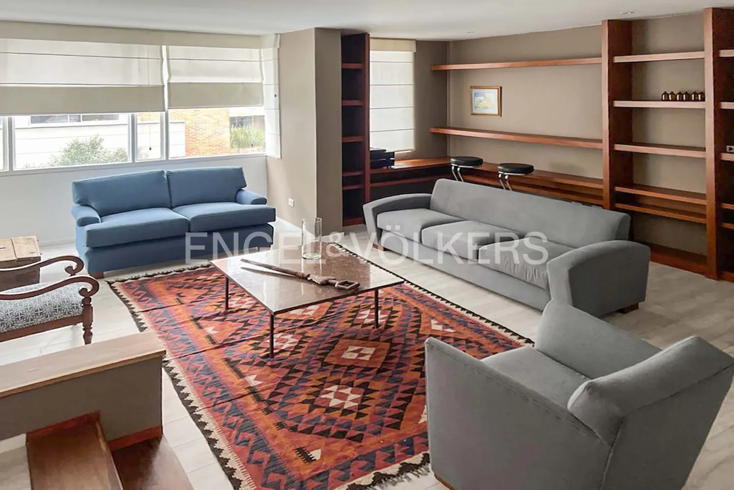 Living room with a blue sofa, grey sofa and armchair, patterned rug, marble coffee table, and wooden built-in shelving.
