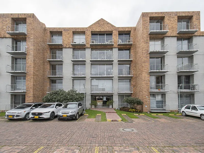 Exterior view of a multi-story apartment building with brick and gray concrete facade, balconies, and parked cars.