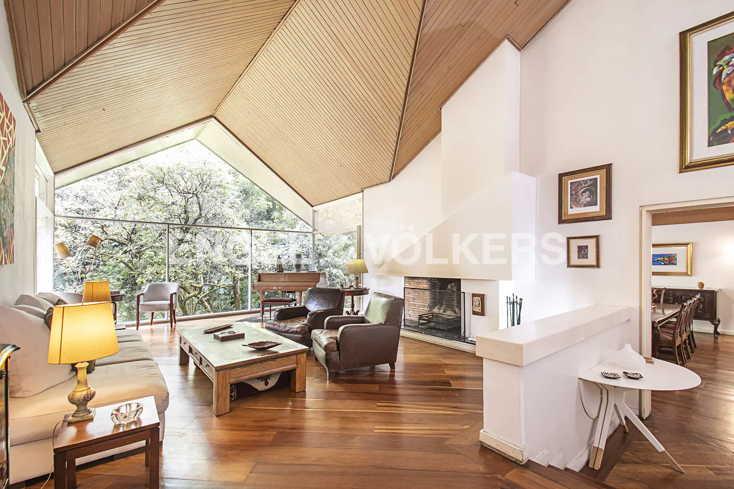 A bright living room with wood floors, a high wood ceiling, a fireplace, and a large window with a view of trees.