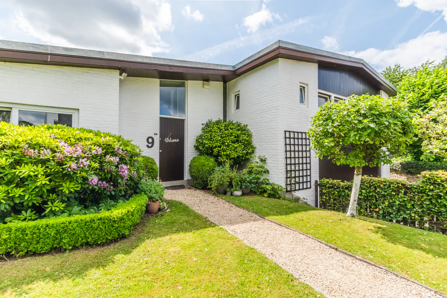 White brick house with a gravel path leading to a dark front door, surrounded by lush green landscaping and a blue sky.