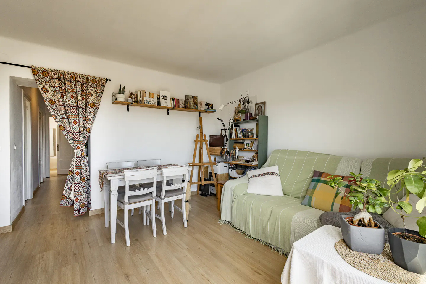 Bright living room with a white table and chairs, a sofa with a green blanket, and a wooden easel. Bookshelves are on the wall.