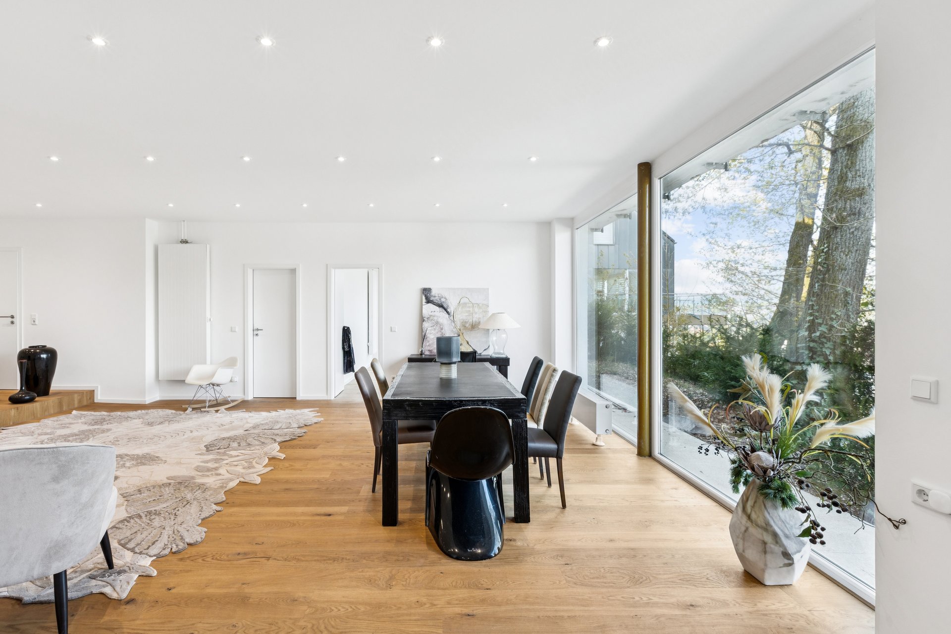 Modern dining room with a black table, dark chairs, hardwood floors, and a large window overlooking trees. [AI-generated text]