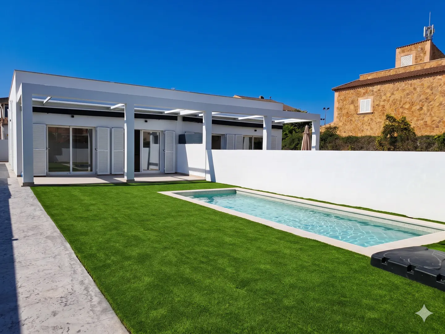 Modern white house with a rectangular pool and green lawn under a clear blue sky. A stone building is visible in the background.