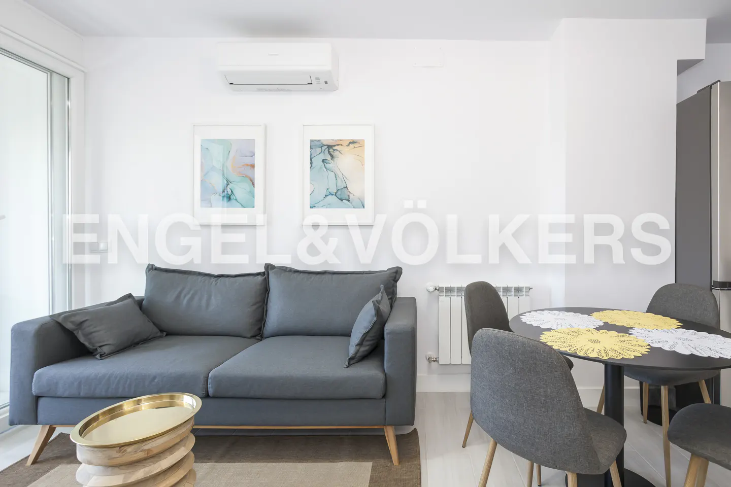 Modern living room with a grey sofa, round dining table with grey chairs, and abstract wall art in a bright white space.