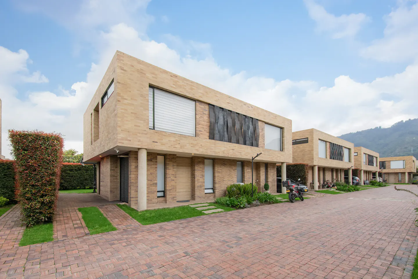 Modern townhouses with brick facades and large windows line a brick driveway under a cloudy sky. A motorcycle is parked near one unit.