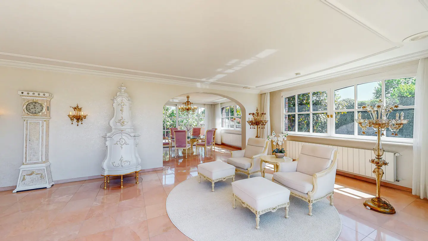 Elegant living room with white armchairs, ottomans, and a round rug. A grandfather clock and decorative fireplace stand against the wall. Large windows offer a view of lush greenery.