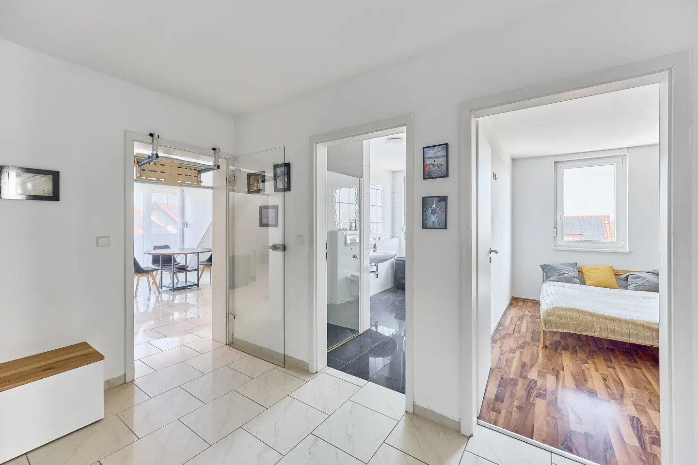White hallway with tiled floor leading to a bathroom and a bedroom with a wooden floor and a bed with yellow pillows.