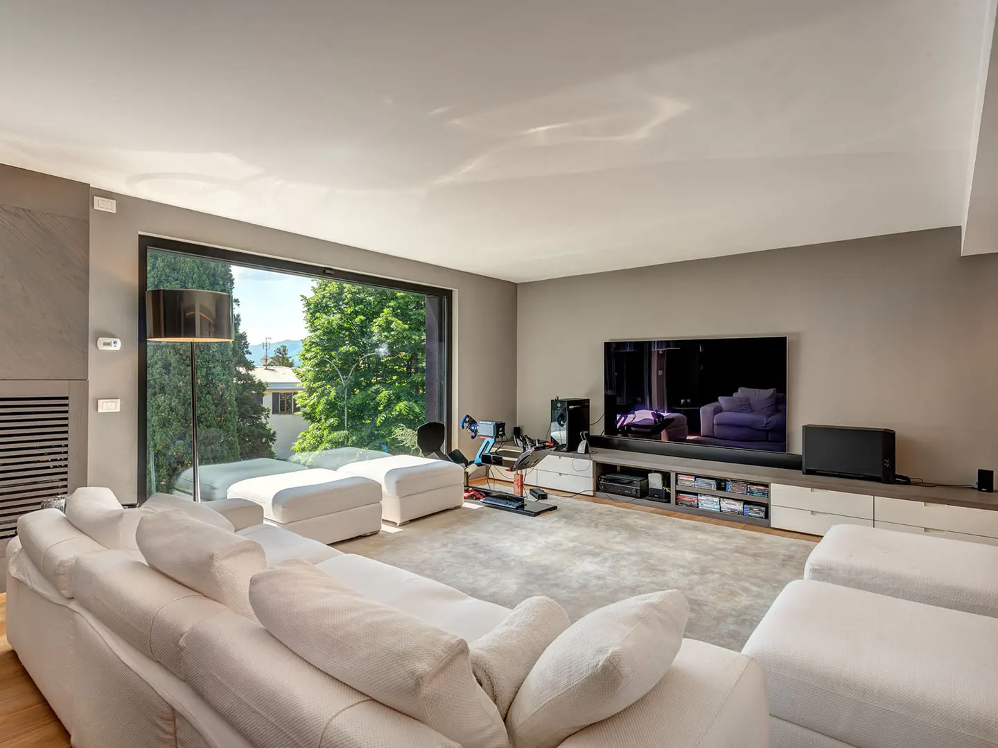 Living room with a large window showing trees and a lake. A white sofa faces a TV and gaming setup.