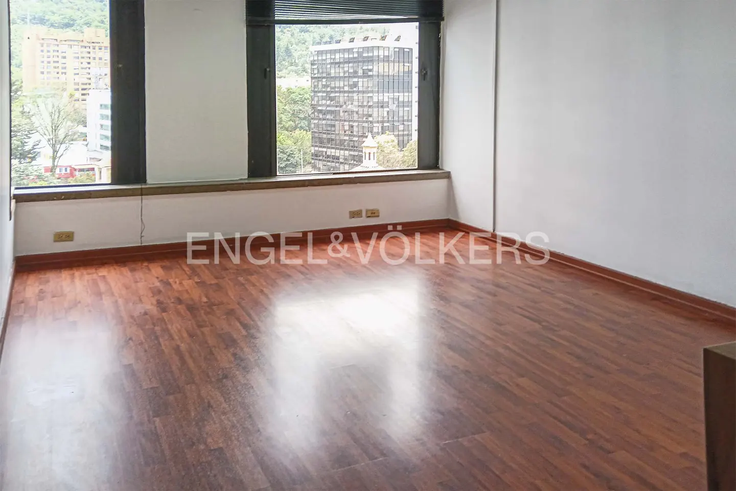 Empty room with wood floors, white walls, and large windows overlooking a city.