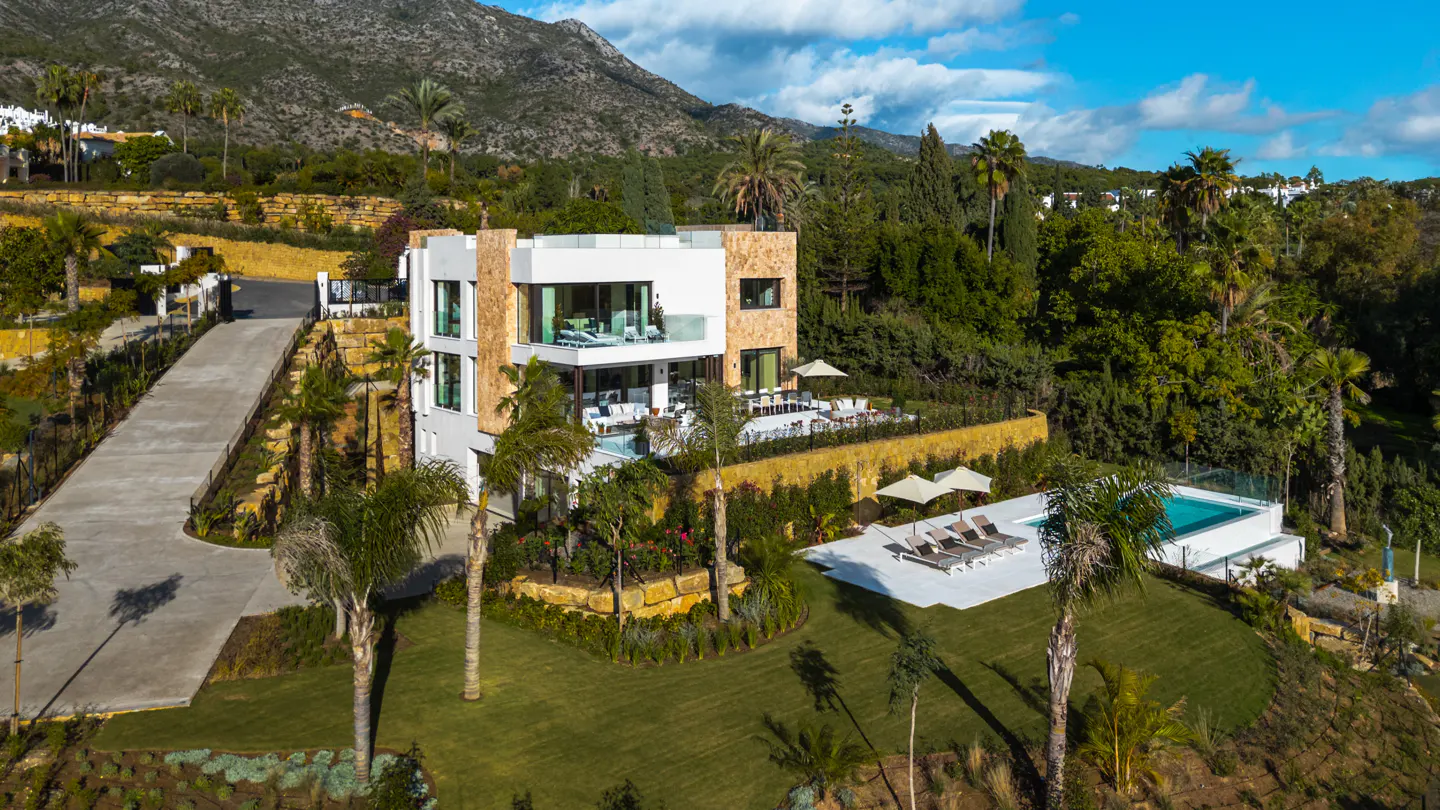 Aerial view of a modern, multi-story white and tan house with a pool, lounge chairs, and lush green landscaping against a mountain backdrop.