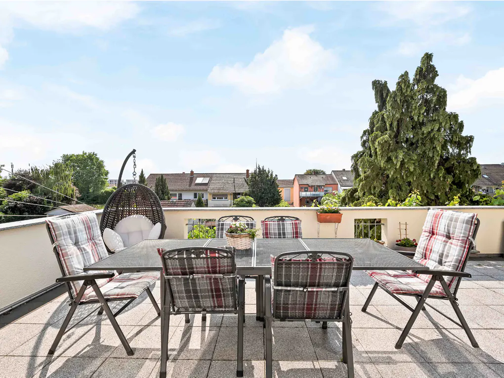Outdoor patio with a metal table, chairs with plaid cushions, and a hanging chair. Trees and houses are in the background.