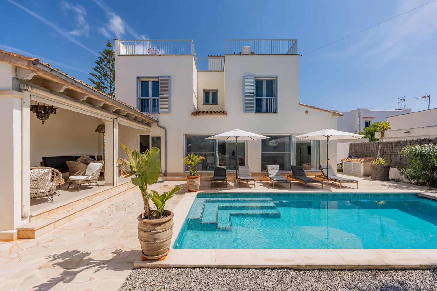 Exterior of a two-story white house with a blue tiled pool, lounge chairs, and umbrellas on a sunny day.