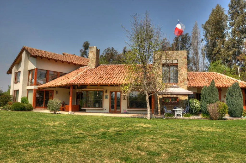 Exterior view of a large, two-story house with a red tile roof and a green lawn. A Chilean flag flies on top of the house.